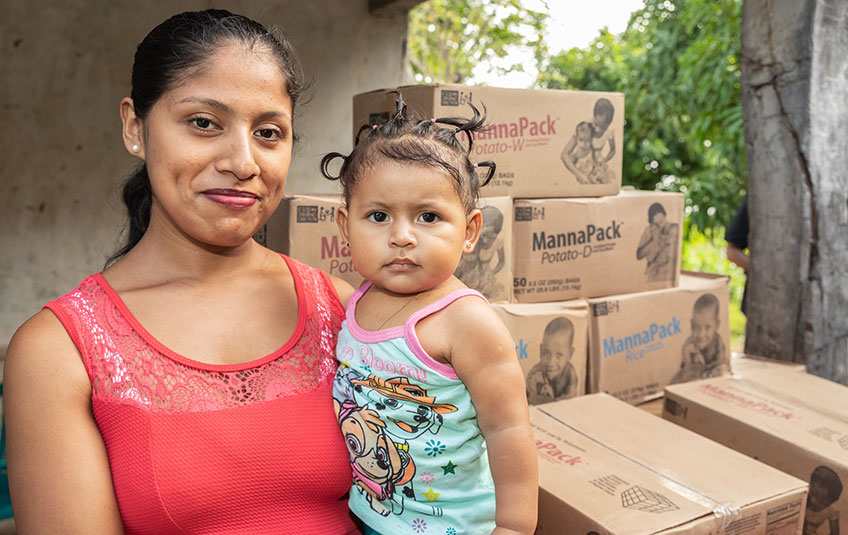 a mother holding her daughter in front of boxes of FMSC'S MannaPack meals